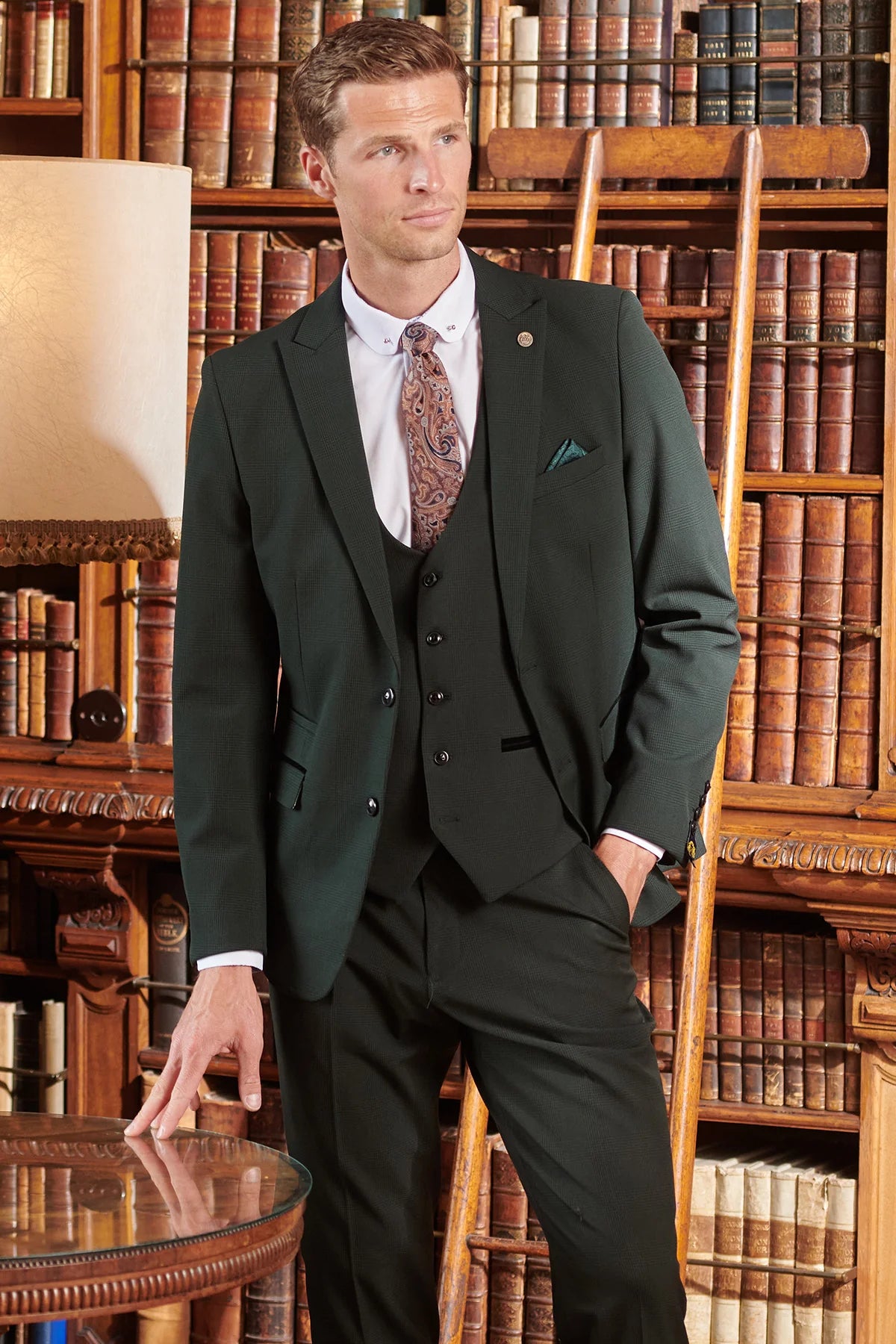 A man wearing an olive green check three piece suit with a blazer, waistcoat, and trousers, standing in a room with bookshelves in the background.
