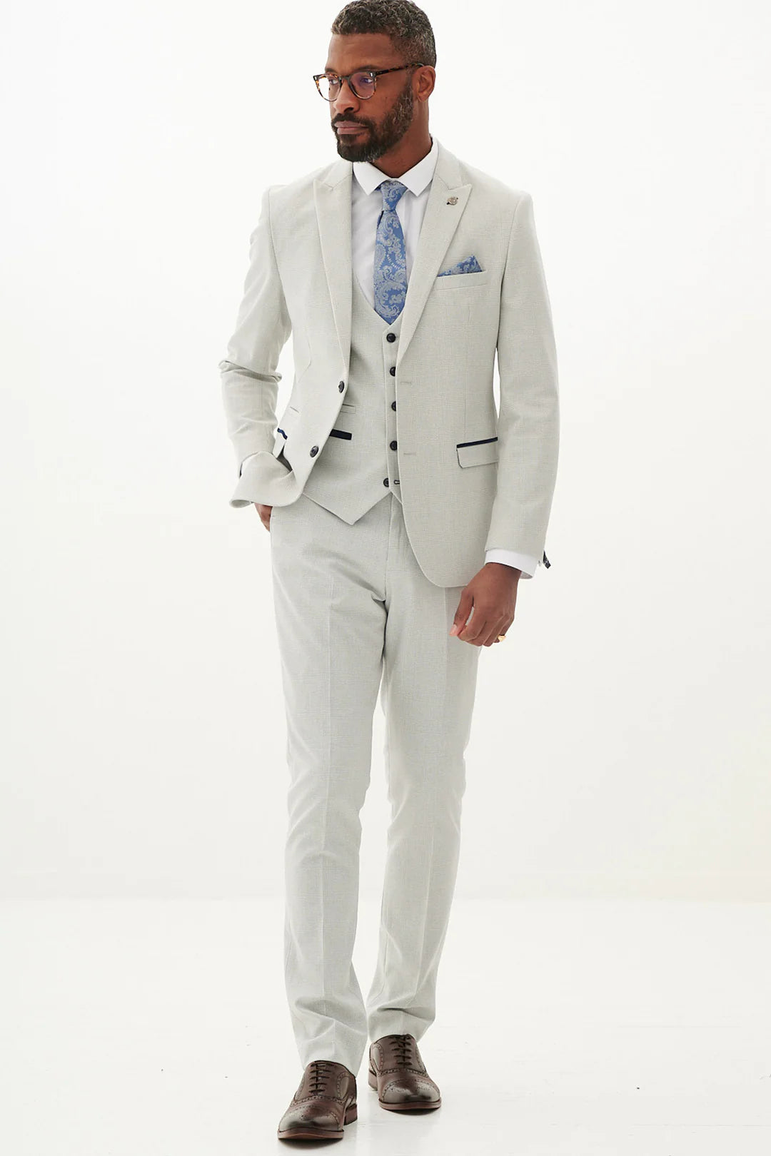 A man standing against a white background, wearing a stone check three piece suit with a peak lapel, double-breasted waistcoat, and brown shoes.