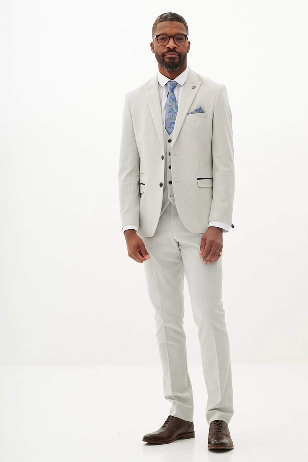 A man standing against a white background, wearing a stone check three piece suit with a peak lapel, double-breasted waistcoat, and brown shoes.