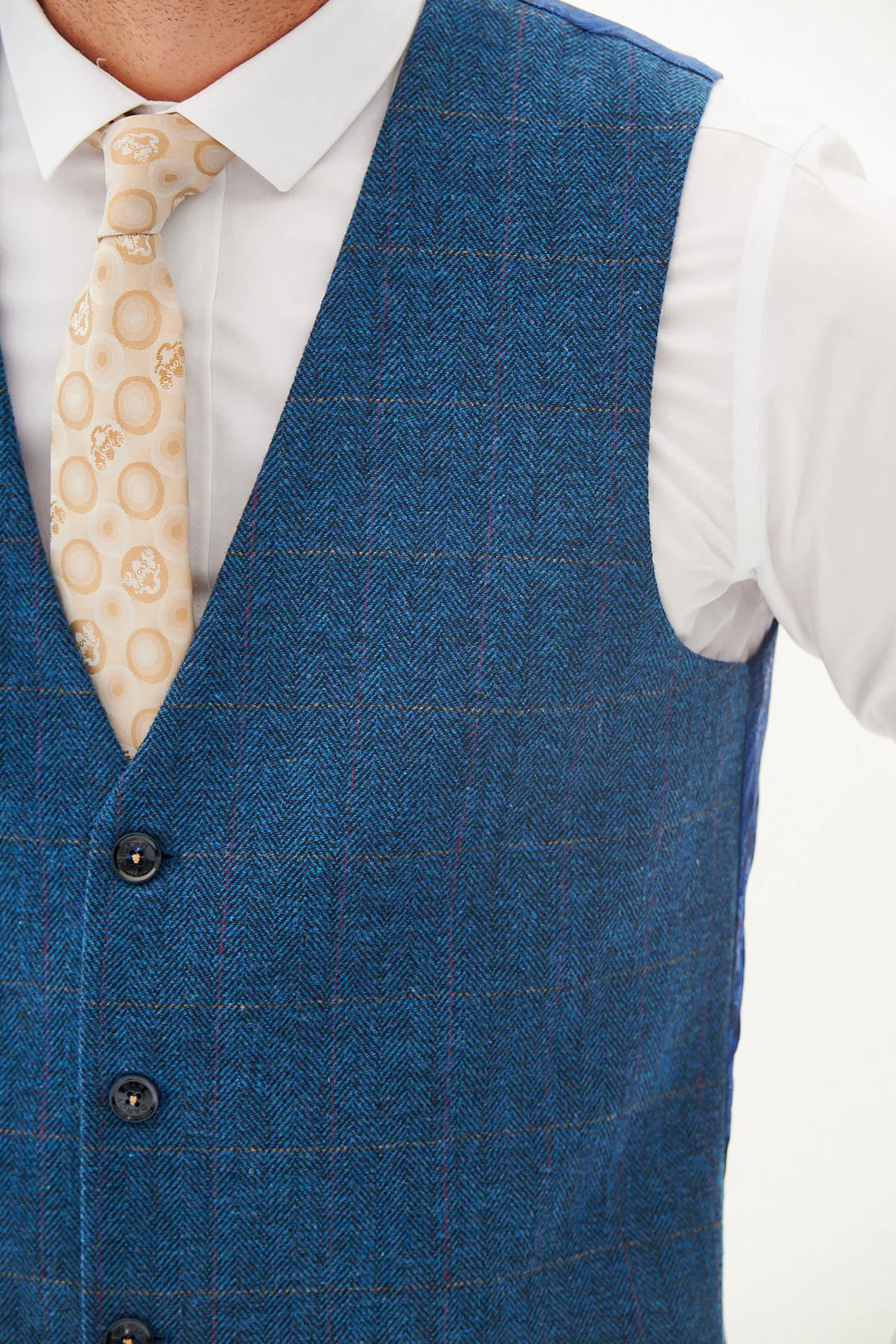 A man wearing a blue tweed check three piece suit with a blue tie and brown shoes.