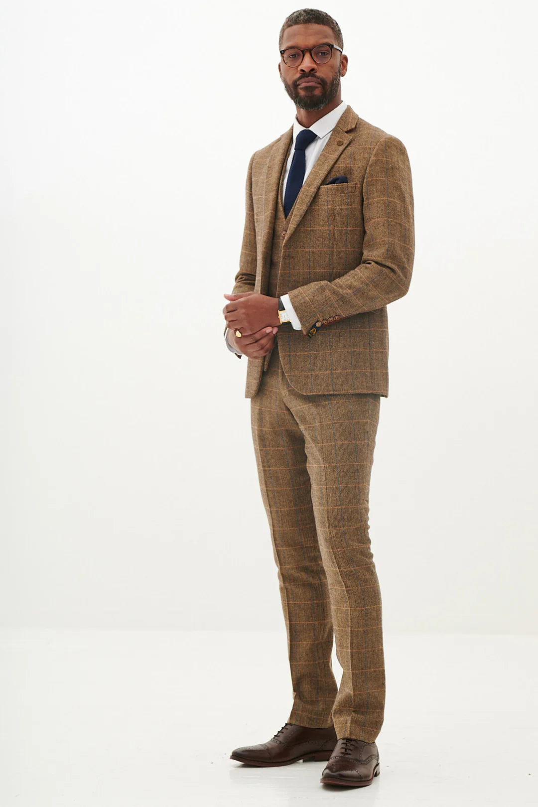 A man wearing a tan tweed check three piece suit with a blue tie and brown shoes.