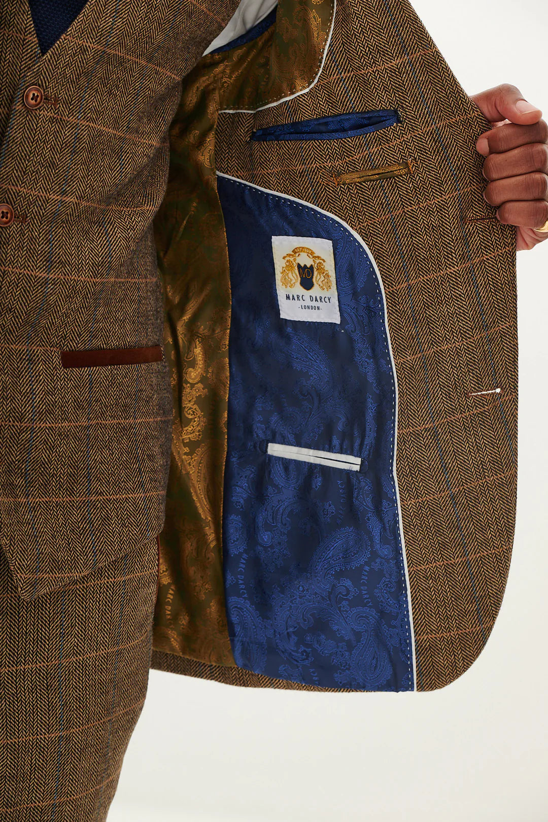 A man wearing a tan tweed check three piece suit with a blue tie and brown shoes.