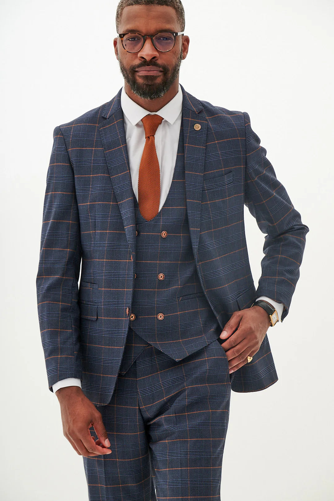 A man standing in a stadium seating area wearing a navy blazer with a check pattern, paired with a white shirt and a rust orange tie.
