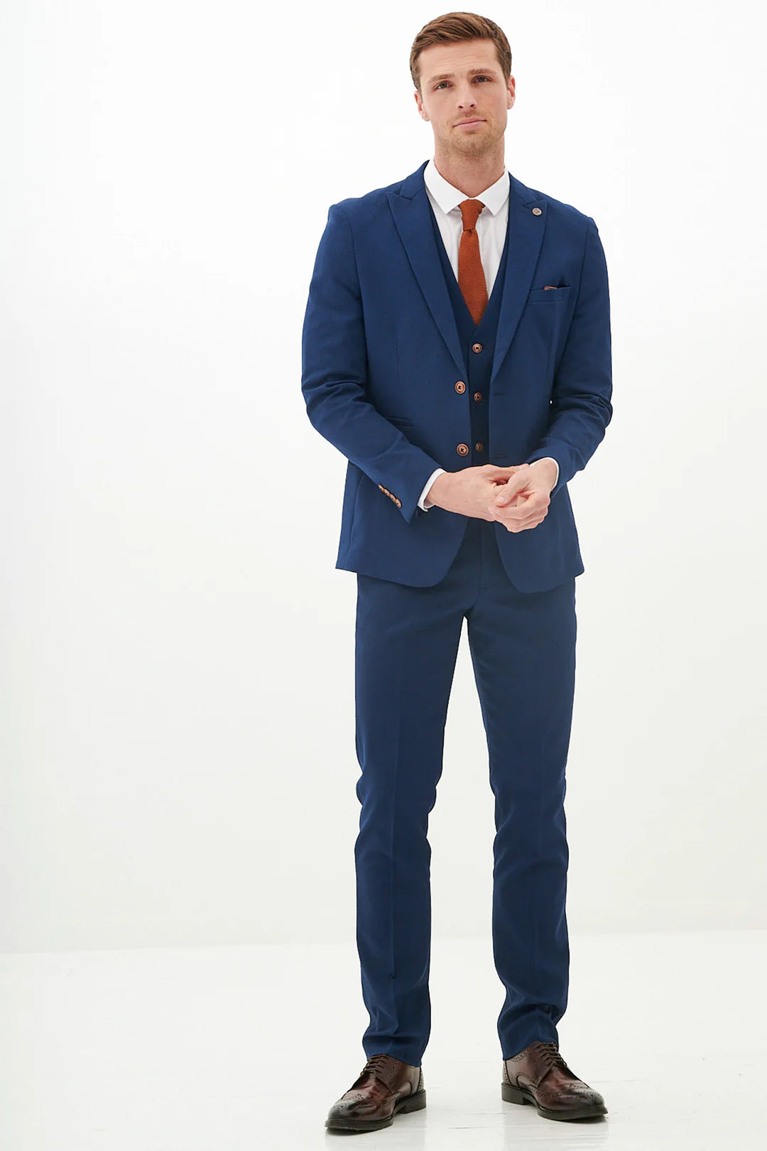 A man standing against a white background, wearing a royal blue three-piece suit with a peak lapel, a white shirt, and brown shoes. He has a pocket square that matches the suit.