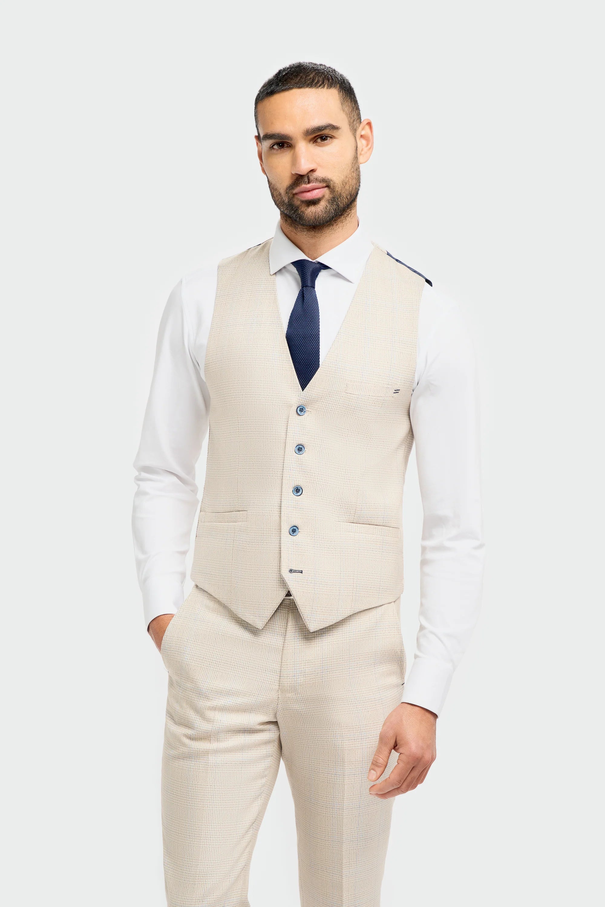A male model wearing a beige check three piece suit with a classic blazer, waistcoat, and trousers, paired with a blue tie and brown shoes.