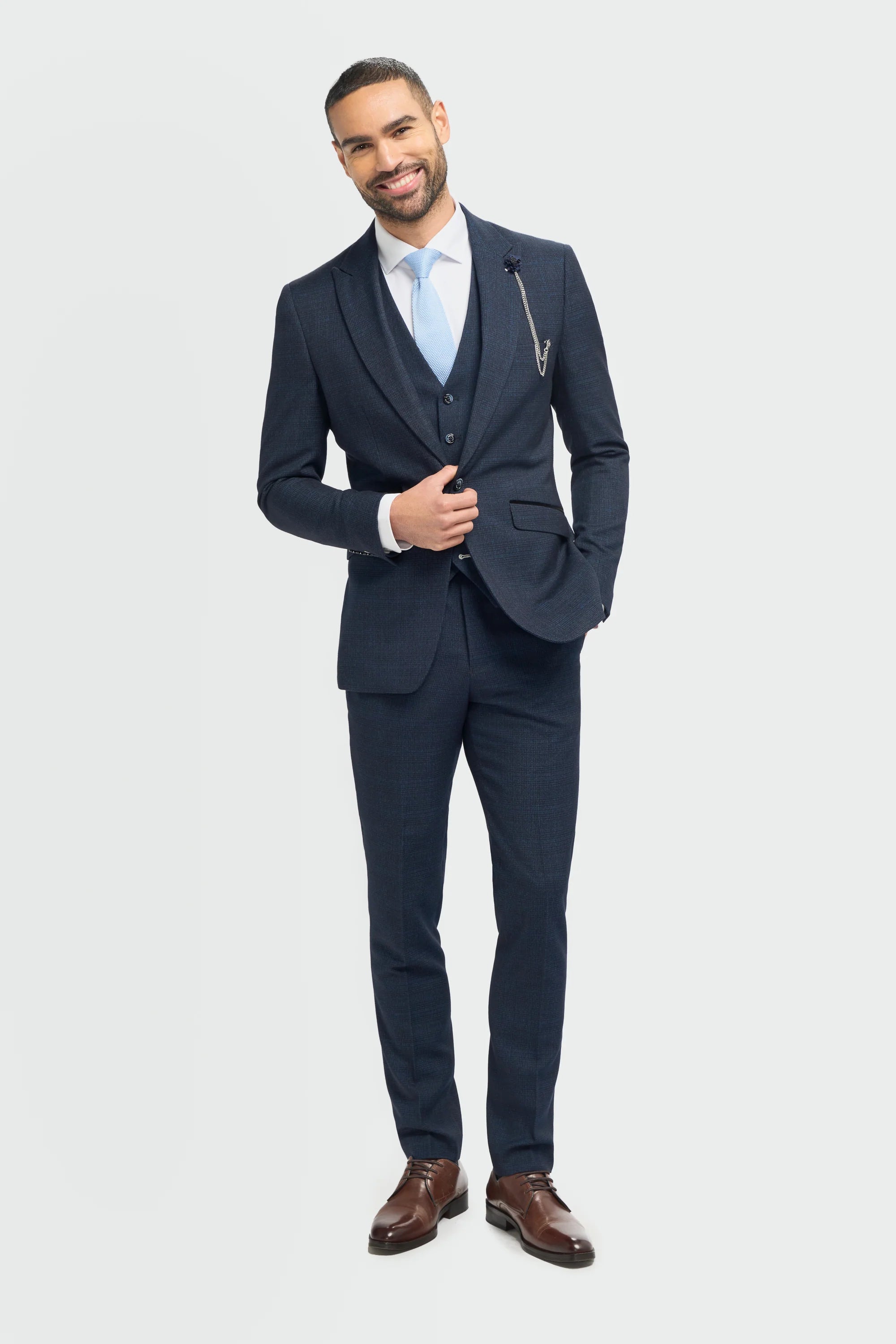A male model wearing a House Of Cavani Caridi Navy Three Piece Suit with a deep navy color, tailored fit, and a regular length. The model is also wearing a white shirt and brown shoes.