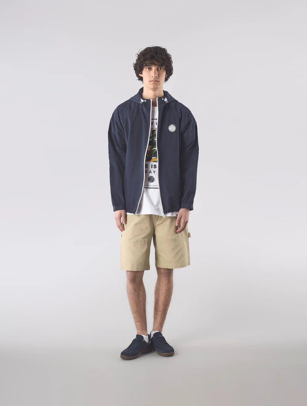 Man wearing a navy jacket, white t-shirt with graphic design, beige shorts, and dark shoes on a light gray background