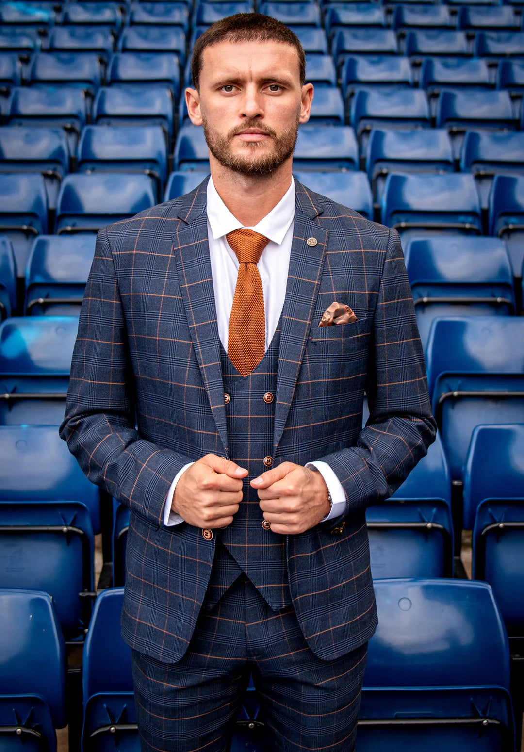 A man standing in a stadium seating area wearing a navy blazer with a check pattern, paired with a white shirt and a rust orange tie.