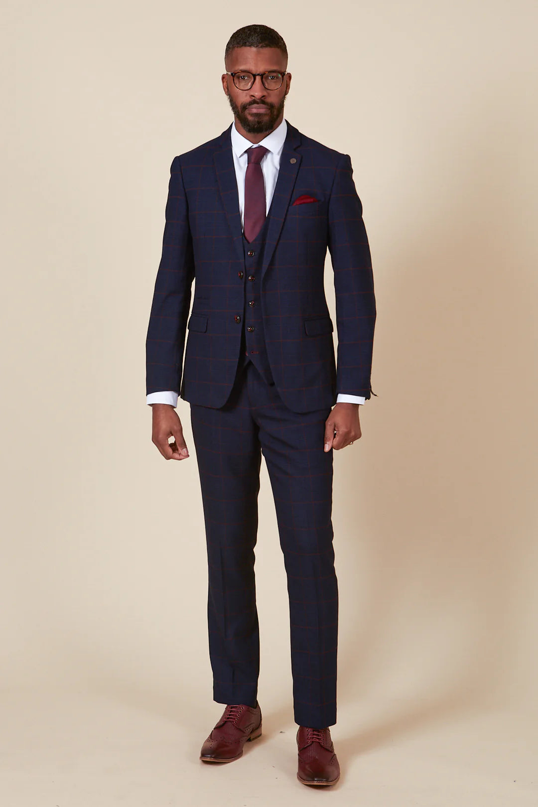 A man modeling a navy three piece suit with a deep burgundy check, consisting of a blazer, waistcoat, and trousers, paired with a red tie and brown shoes.