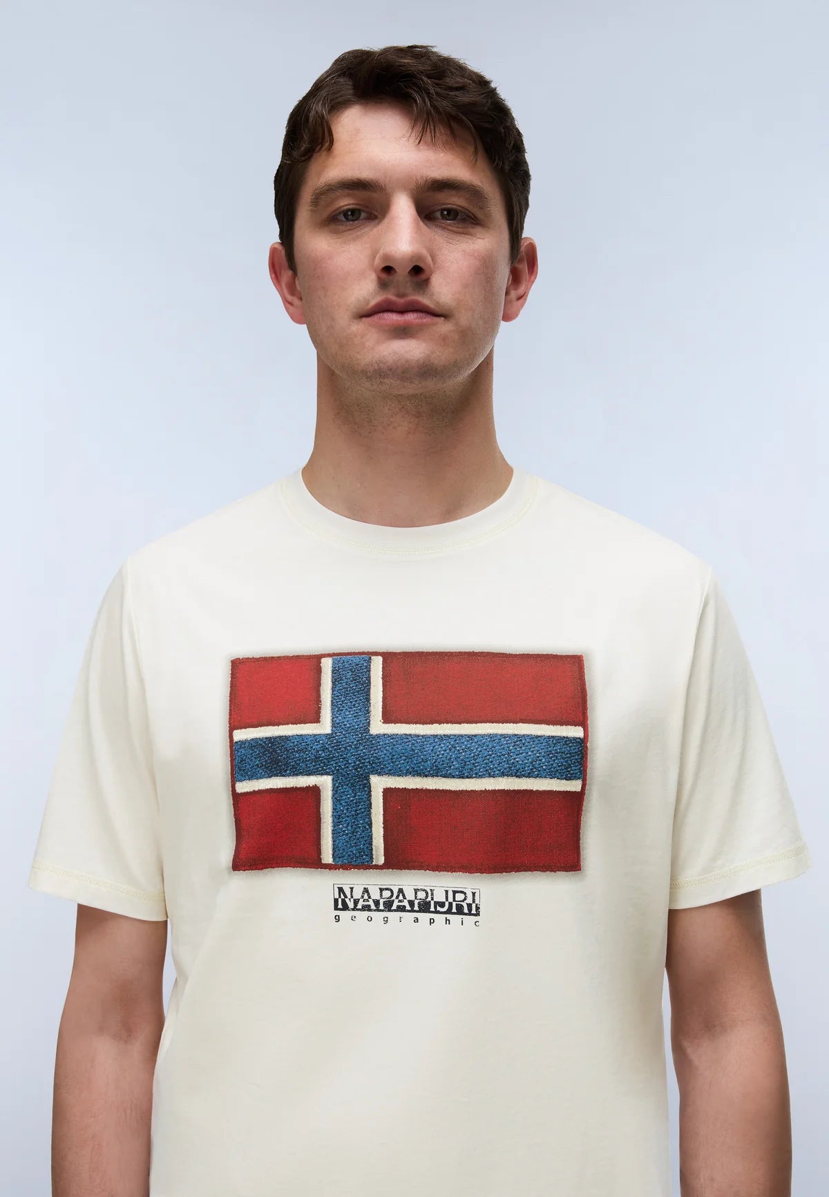 A man wearing a cream-colored t-shirt with a distressed graphic of the Norwegian flag and the brand name 'NORDAGARI' printed below it, set against a light gray studio background.