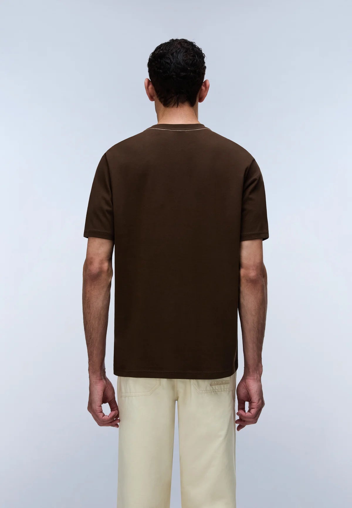 A man viewed from the back wearing a dark brown short-sleeve t-shirt and light beige trousers, standing against a light gray studio background.