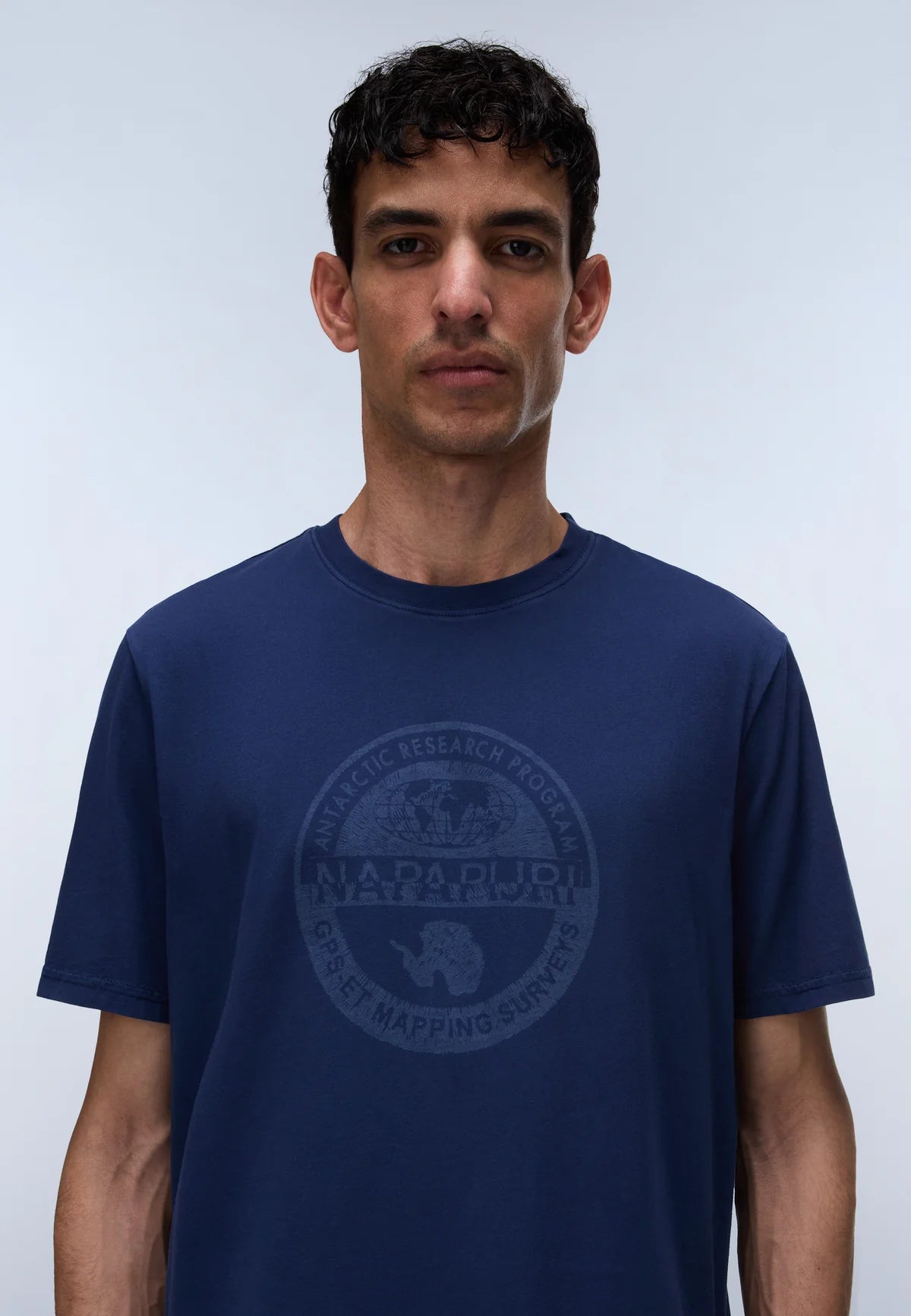 A man wearing a navy blue crewneck t-shirt with a dark gray circular graphic on the chest featuring a polar bear and the text 'NARNUPIU ARCTIC RESEARCH FOUNDATION' and 'ARCTIC MAPPING SURVIVOR'.