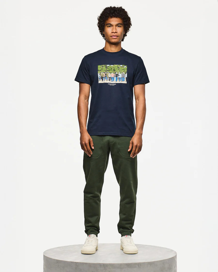 Man wearing a navy t-shirt with a graphic design and green pants on a white background