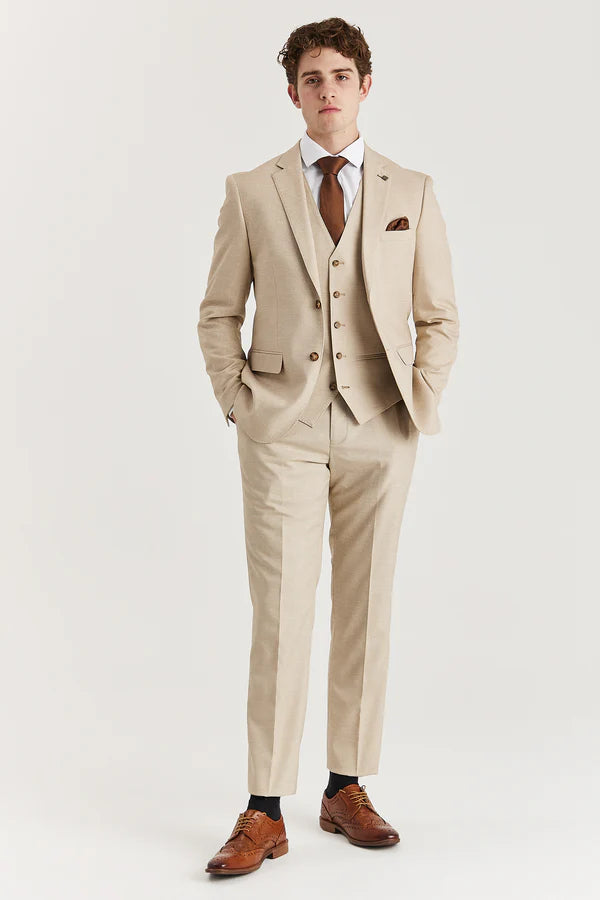 A man wearing a beige three-piece suit with a jacket, waistcoat, and trousers, paired with a white shirt and brown shoes.