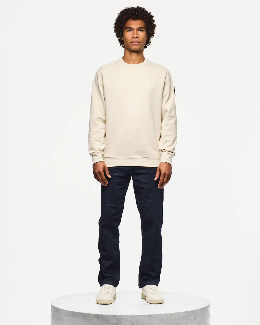 Man wearing a beige sweatshirt and dark jeans on a white background