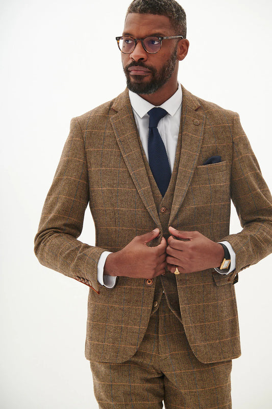 A man wearing a tan tweed check three piece suit with a blue tie and brown shoes.