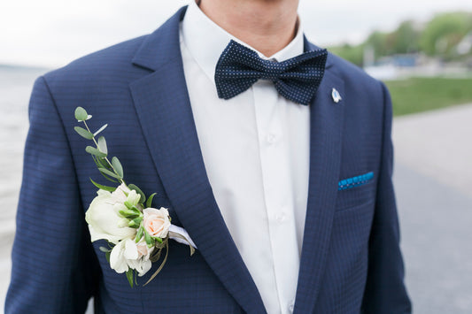 Man wearing a navy suit with a white shirt, bow tie, and floral boutonniere outdoors.