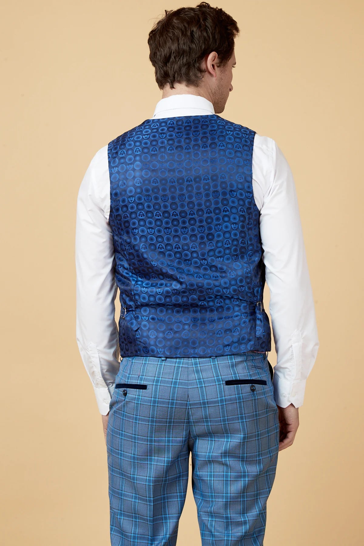 Man wearing a blue patterned vest over a white shirt and blue plaid pants on a beige background