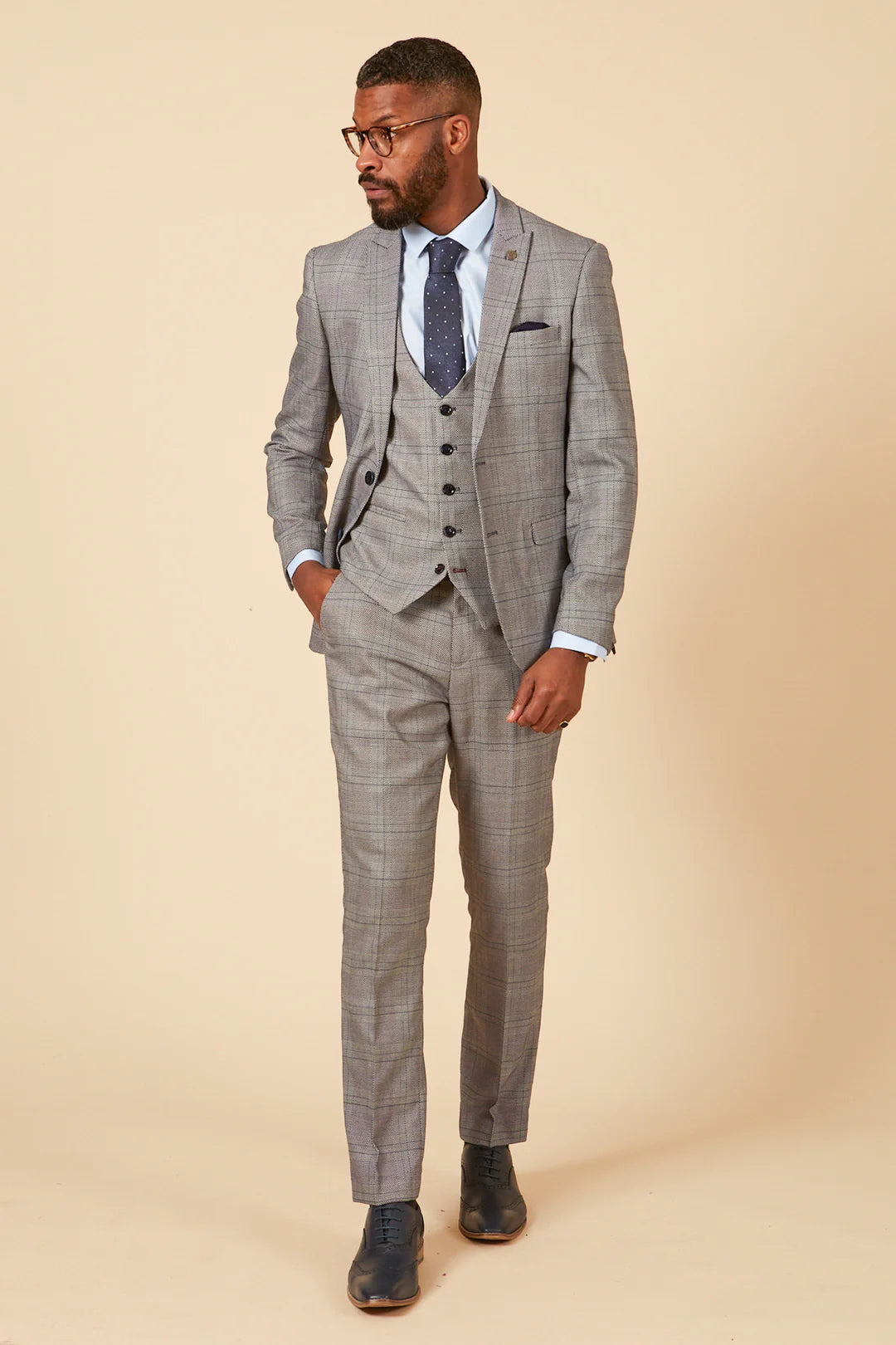 A man wearing a grey check three piece suit with a peak lapel, double-breasted blazer, and a waistcoat. He has a blue tie and the suit has a subtle check pattern.