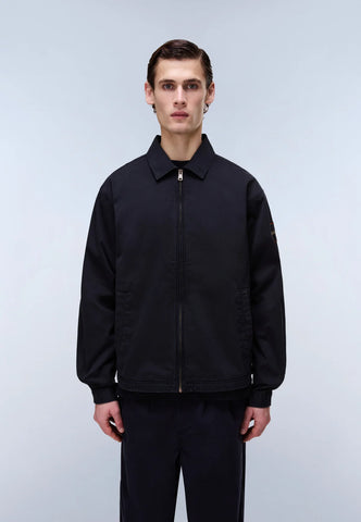 A man wearing a black zip-up jacket with a collar and black pants, standing against a light gray studio background.
