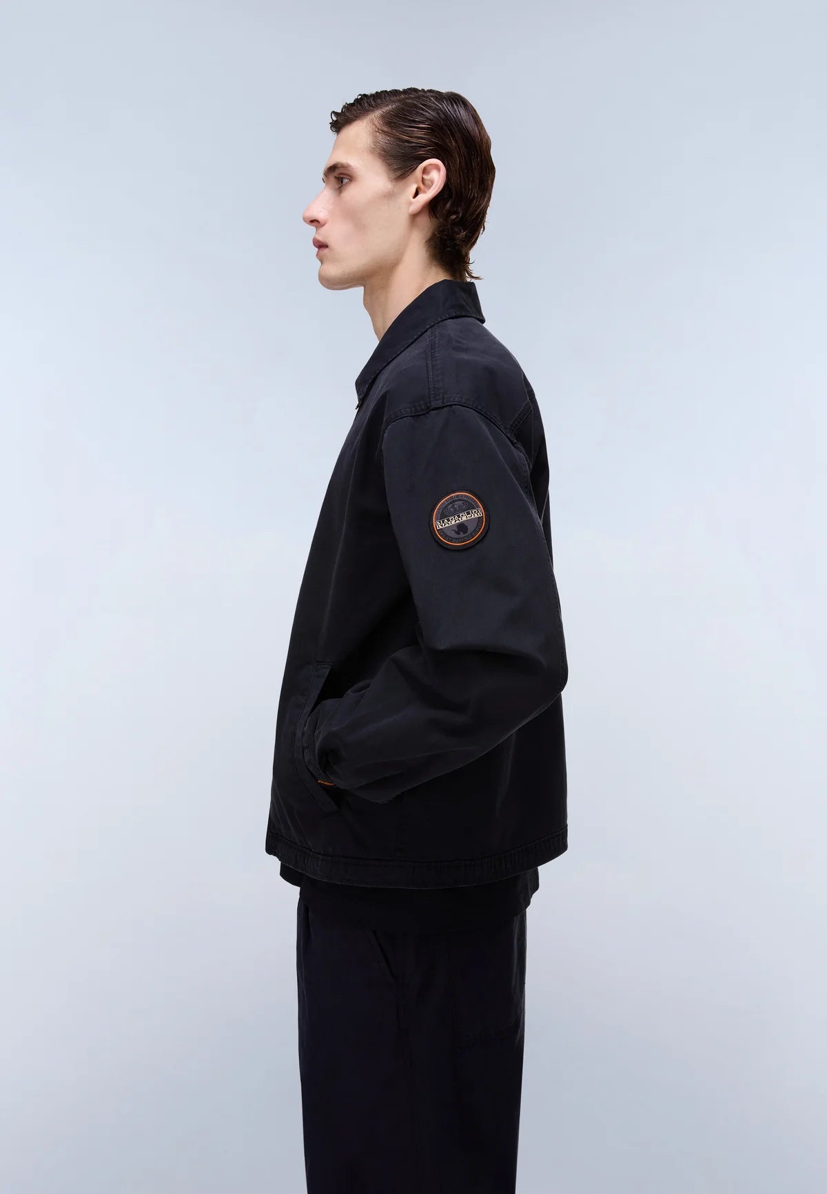 A man in profile wearing a black utility jacket with a circular patch on the upper left sleeve, paired with black trousers, against a light gray studio background.
