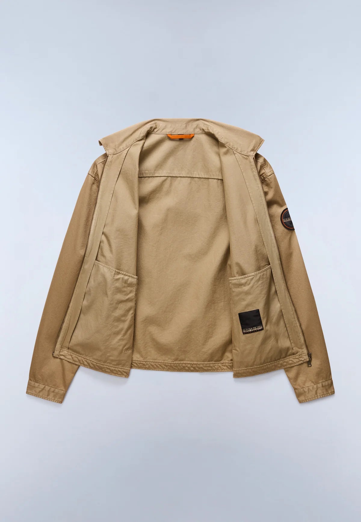 A tan utility jacket shown from the back, featuring a hood, large patch pockets, and a small black brand patch on the lower back and a circular sleeve patch, photographed on a light gray background.