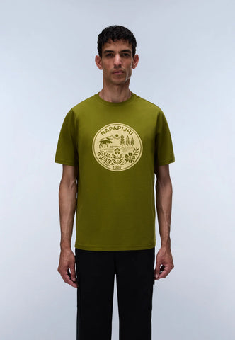 A man wearing a short-sleeve olive green t-shirt with a circular tan graphic on the chest, paired with black pants, standing against a light gray studio background.