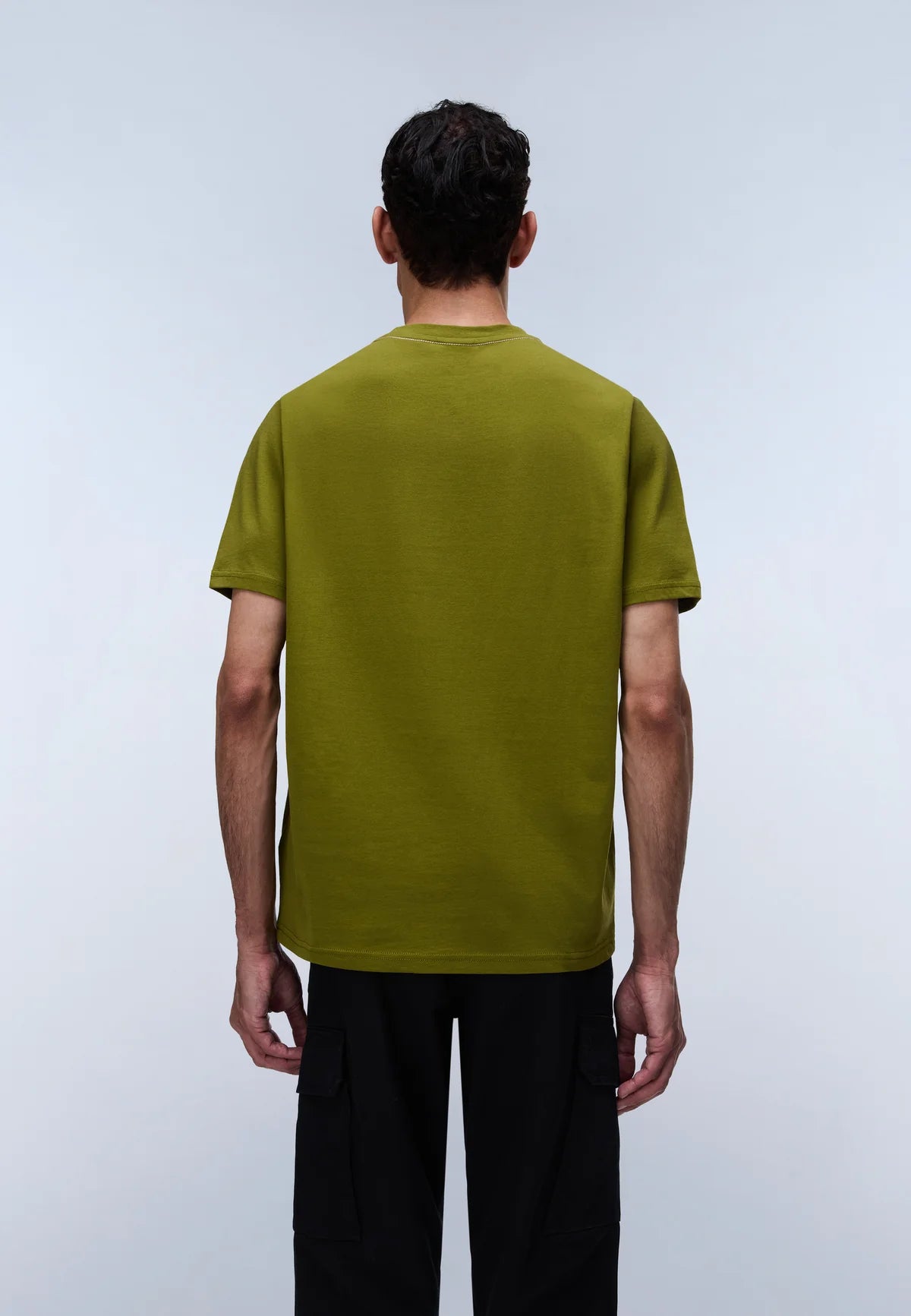A man viewed from behind wearing a short-sleeve olive green t-shirt and black pants, standing against a light gray studio background.