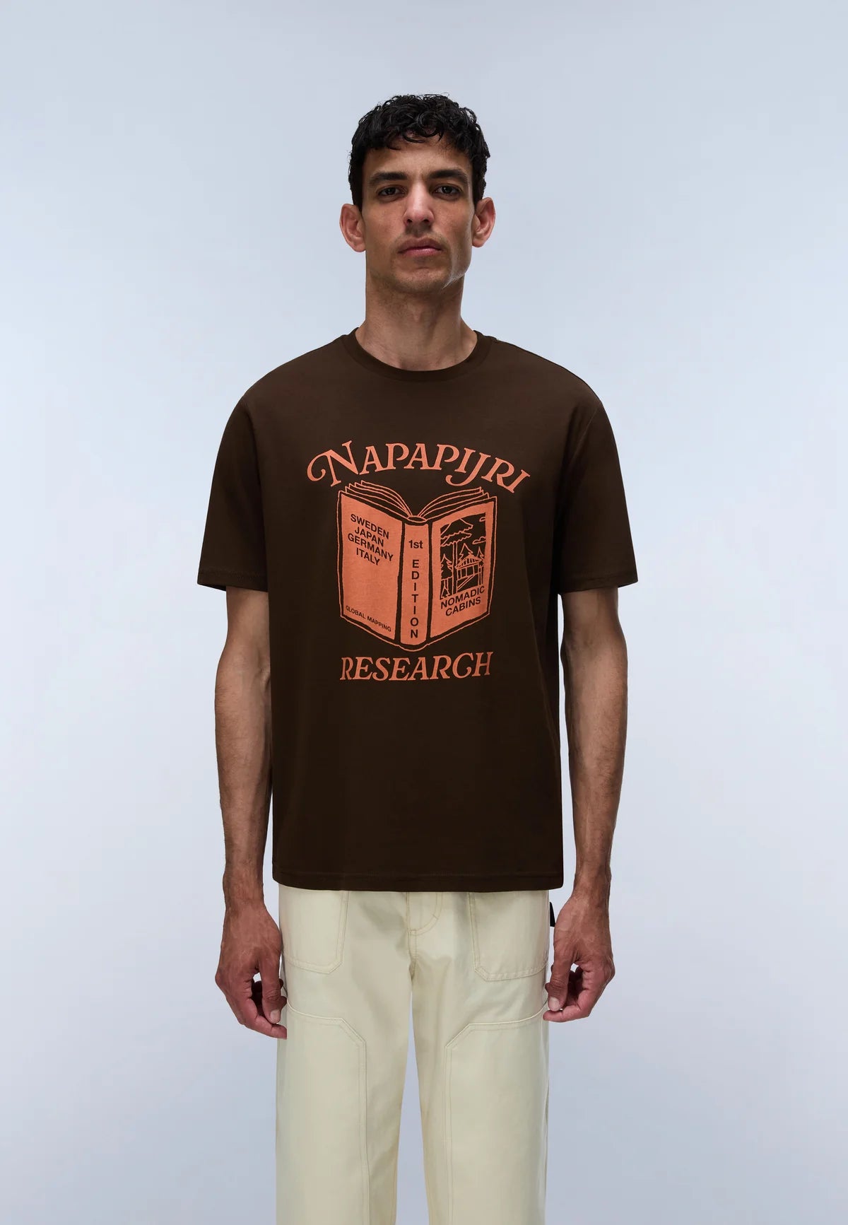 A man wearing a dark brown short-sleeve t-shirt with a graphic print of an open book and the words 'NAPAPIJRI RESEARCH' in orange, paired with light beige trousers, standing against a light gray studio background.