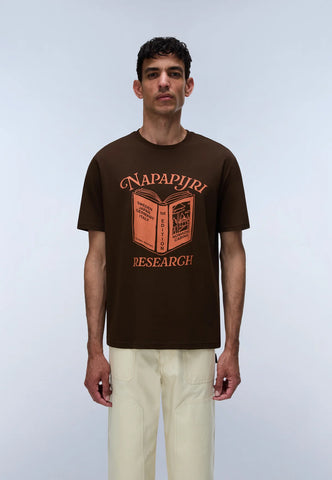 A man wearing a dark brown short-sleeve t-shirt with a graphic print of an open book and the words 'NAPAPIJRI RESEARCH' in orange, paired with light beige trousers, standing against a light gray studio background.