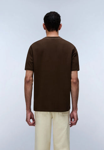 A man viewed from the back wearing a dark brown short-sleeve t-shirt and light beige trousers, standing against a light gray studio background.