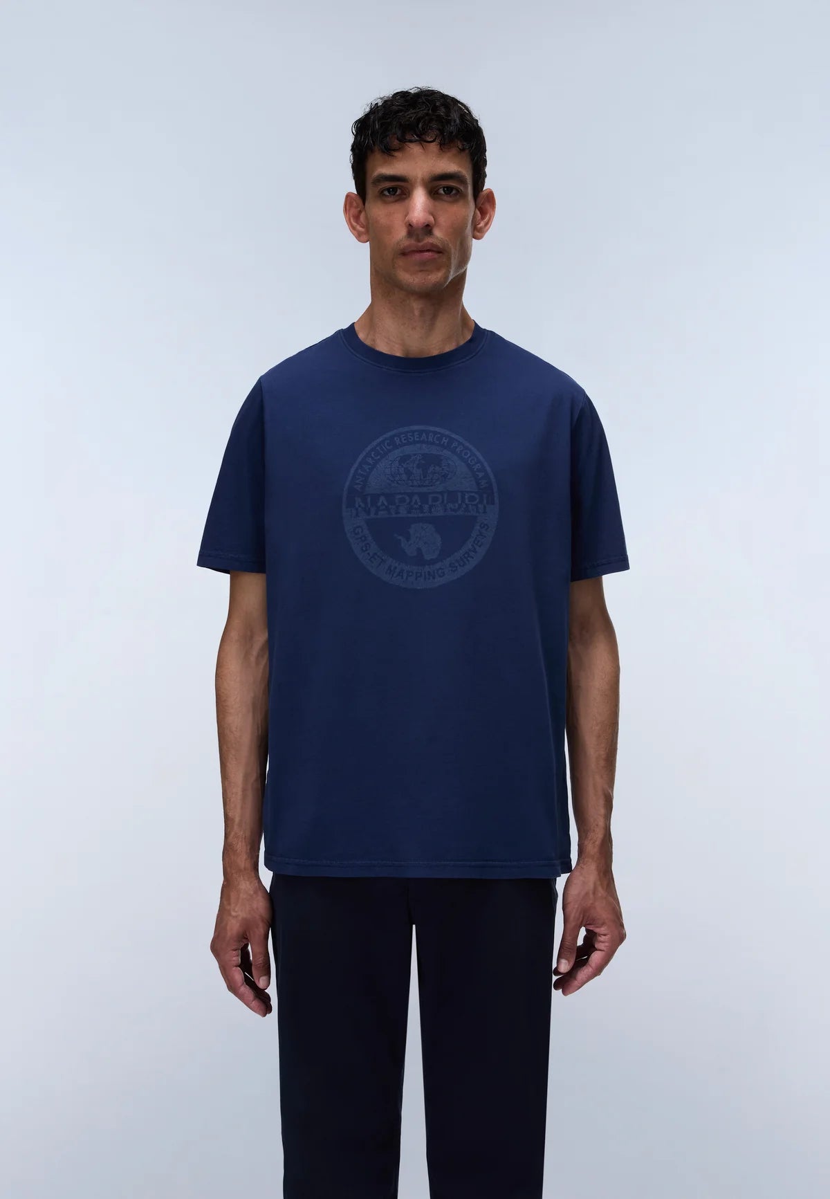 A man wearing a navy blue short-sleeve t-shirt with a tonal circular graphic on the chest, paired with dark pants, standing against a light gray studio background.
