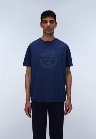 A man wearing a navy blue short-sleeve t-shirt with a tonal circular graphic on the chest, paired with dark pants, standing against a light gray studio background.