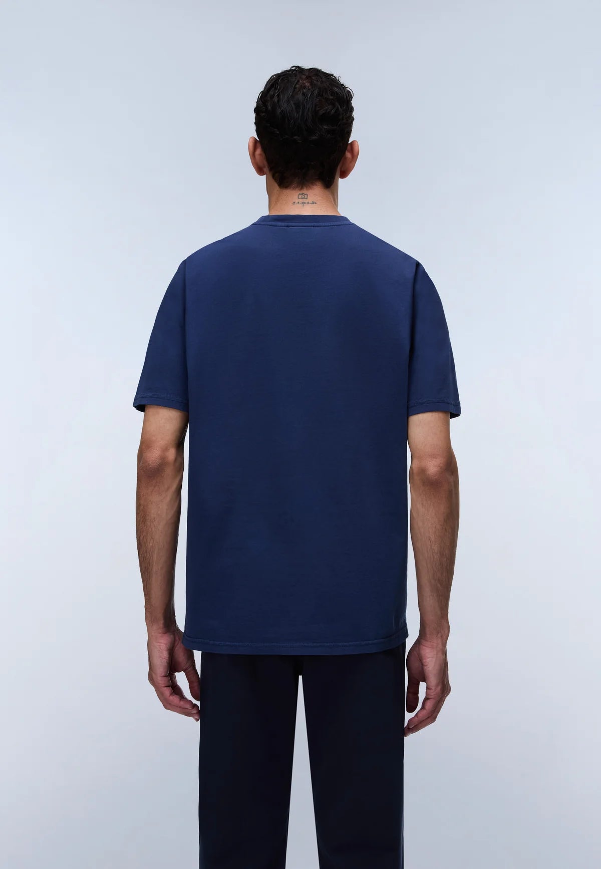 A man viewed from the back wearing a dark navy blue short-sleeve t-shirt and dark pants, standing against a light gray studio background.