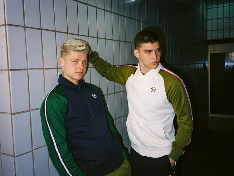 Two young men posing against a tiled wall.