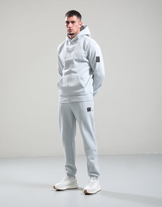 Man wearing a light gray hoodie and pants with a logo on a plain background