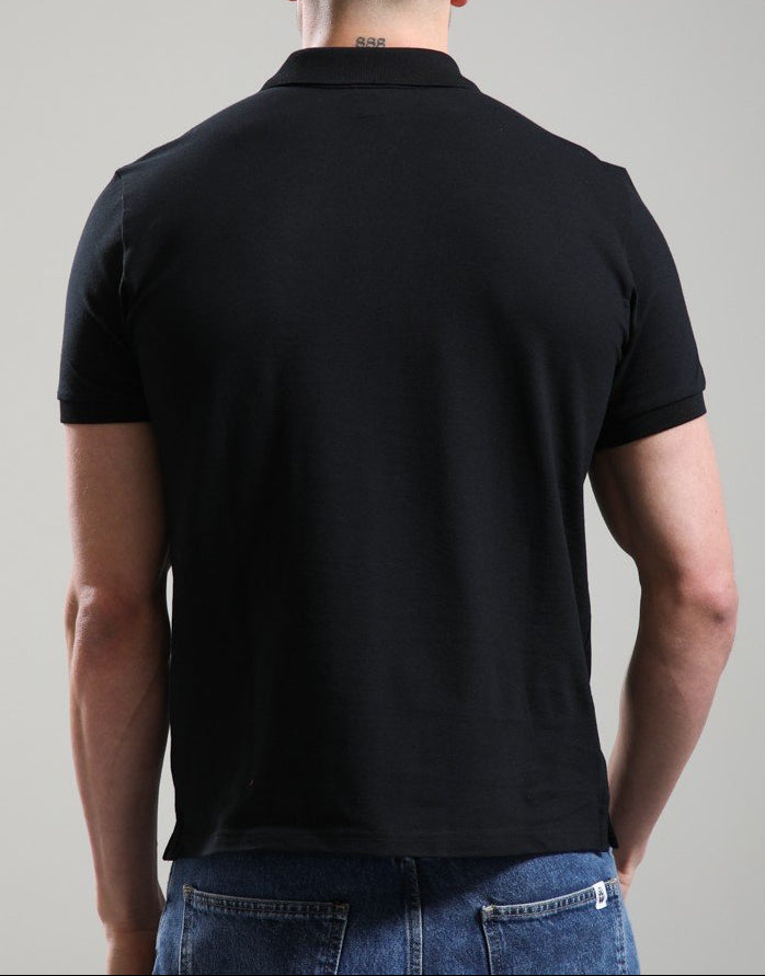 A man viewed from behind wearing a plain black short-sleeve polo shirt and blue jeans, standing against a neutral light gray studio background.
