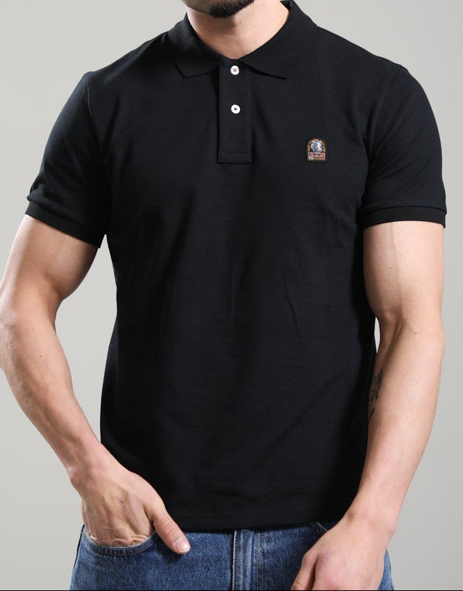 A man wearing a black short-sleeve polo shirt with a small embroidered logo on the left chest, paired with blue jeans, photographed against a solid gray studio background.